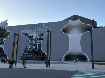 581949879_855000737053866_1824881039060617293_n Modern gray building with white curved arches and a glass entrance, with people walking nearby and a striped crosswalk in the foreground.