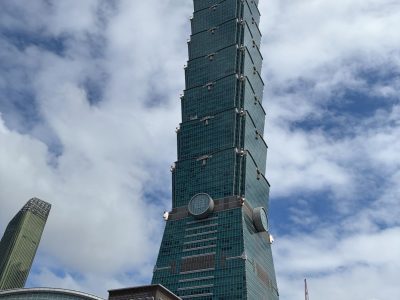 619332903_907610481792891_7558720153520244161_n Taipei 101 skyscraper with blue-green glass, surrounded by construction cranes and a Chanel storefront at its base.