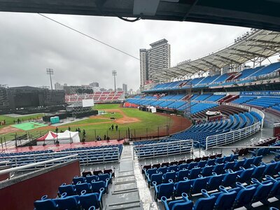 621823634_907605165126756_7612761332693593340_n Wide view of a baseball stadium with a stage setup and scaffolding on the left, surrounded by blue seating and a cloudy sky.